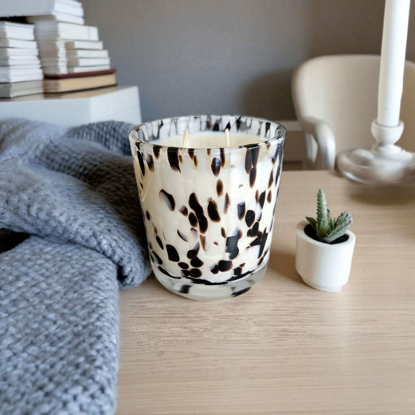 Candle with a dalmatian print pattern on a wooden surface next to a stack of books and a small plant.