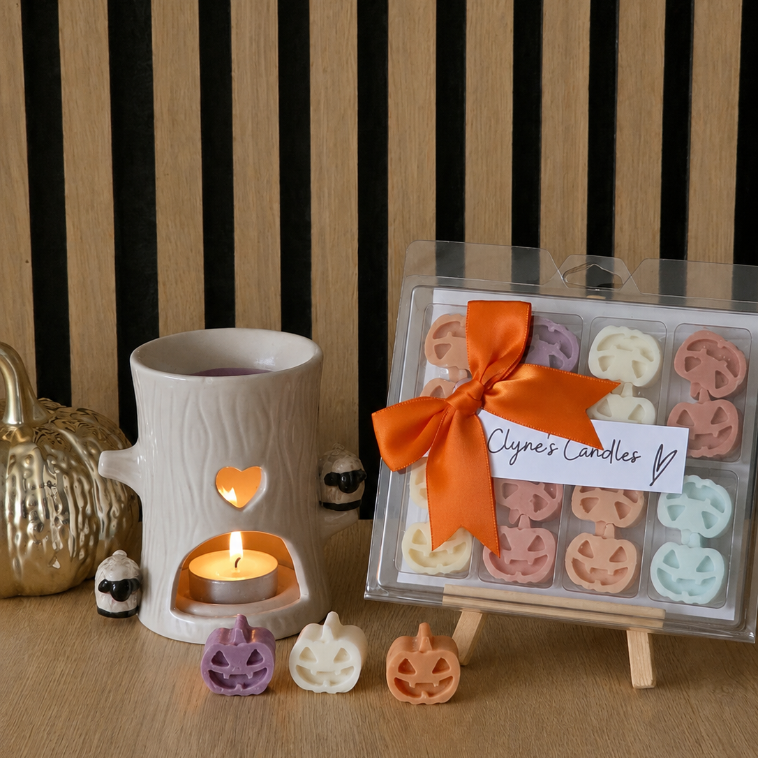 A boxed set of colorful wax melts shaped like pumpkins, tied with an orange ribbon, displayed on a small wooden easel next to a white ceramic candle warmer with a lit tealight candle. The scene is styled with small decorative pumpkins on a wooden surface against a wood-and-black striped background.