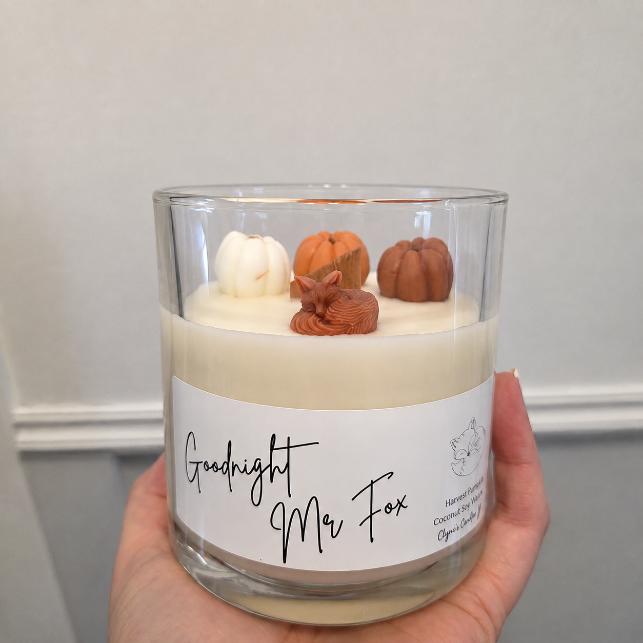 A hand holds a clear glass jar candle with cream-colored wax and decorative autumn-themed toppers, including small pumpkins and a fox figurine. The candle has a white paper label that reads 'Goodnight Mr Fox'.
