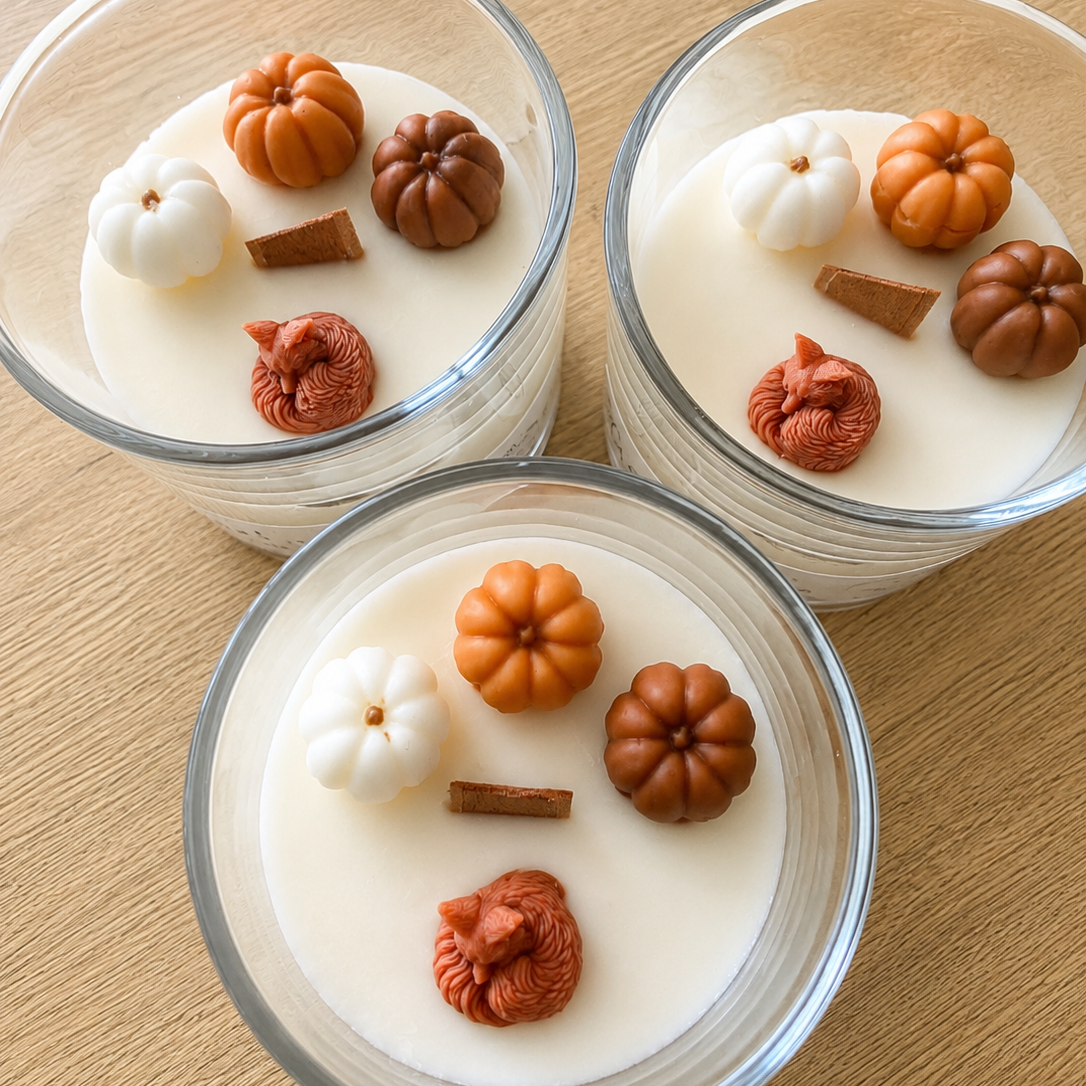 Three clear glass jars filled with creamy white wax or soap, topped with small decorative autumn motifs including orange and brown pumpkins, a white pumpkin, a curled sleeping fox handmade