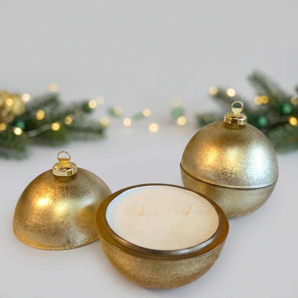 A gold-toned spherical candle holder set on a dark marble countertop, with one lid leaning beside a white wax candle and another lid standing behind it. A white wall outlet is visible above on a beige wall.