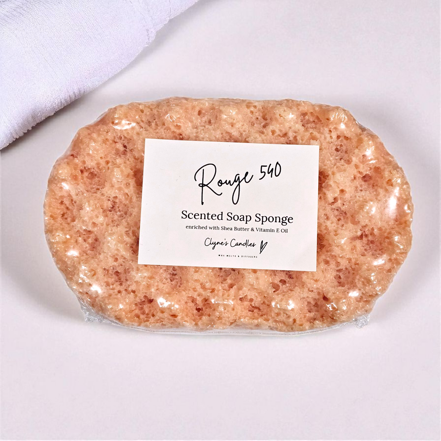 Rouge soap sponge by Clyne’s Candles, a 2-in-1 exfoliating sponge infused with luxurious designer-inspired Rouge fragrance — warm amber, jasmine and woody notes — enriched with shea butter for soft, nourished skin.